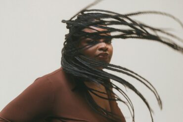 Schwarze Frau, die ihre langen Kynd Hair Braids durch die Luft schwingt und dabei in die Kamera schaut